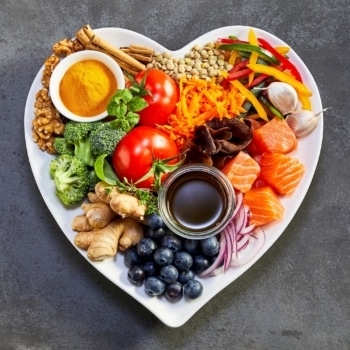 Heart healthy foods, good for lowering cholesterol, in a heart-shaped dish including acai, lentils, soy sauce, ginger, salmon, carrot, tomato, turmeric, cinnamon, walnuts, garlic, peppers, broccoli, basil, onion