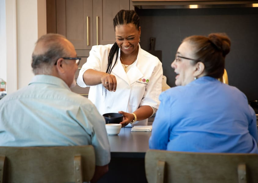 Personal chef seasoning meals in front of elderly couple.