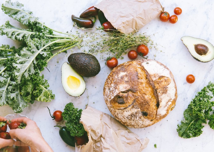 Bread, avocado, tomatoes, kale, and fresh herbs are spread across a white marble countertop. Two hands place fresh strawberries in a small white bowl.