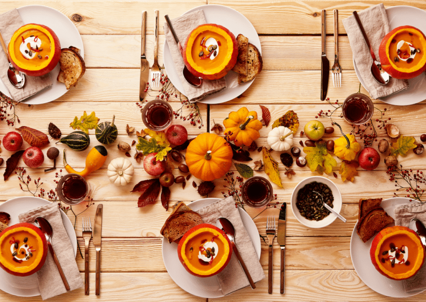 Table with a centerpiece of fall favorites including colorful leaves, pumpkins, squash, and apples, and place settings with linen napkins, gold silverware, and pumpkin soup in small carved pumpkins