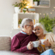 senior couple smiles at each other while cuddling on the couch holding white coffee mugs