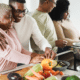 Family cooking a meal together, older woman and young man side-hug and smile fondly while preparing vegetables, other family members cook at the stove in the background
