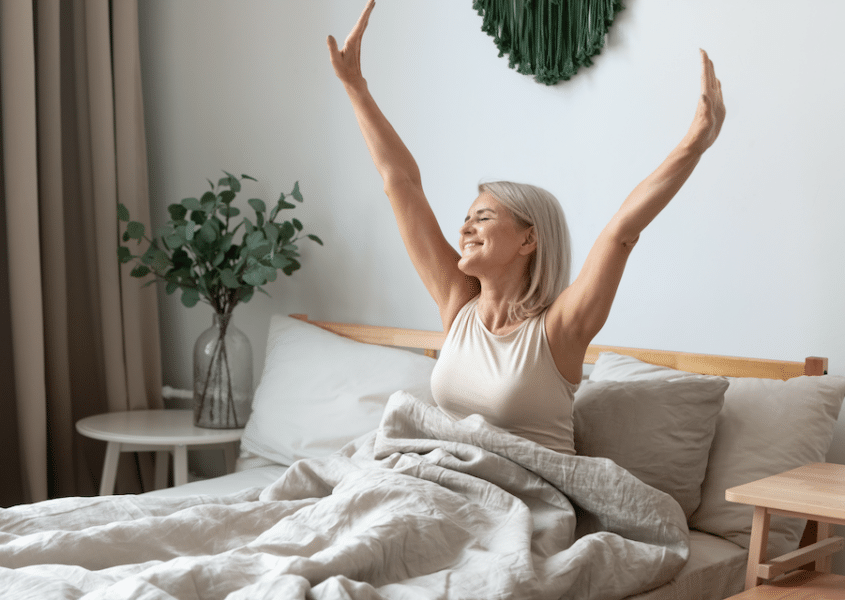 Smiling older woman wakes up happy and refreshed in bed, stretching her arms above her in the morning light after a restful night