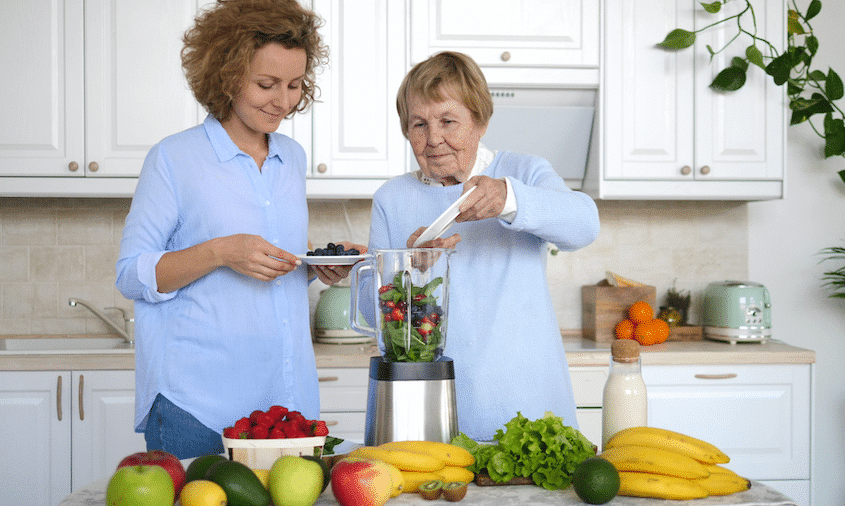 Older adult woman and adult granddaughter make a smoothie with fresh fruits and greens.